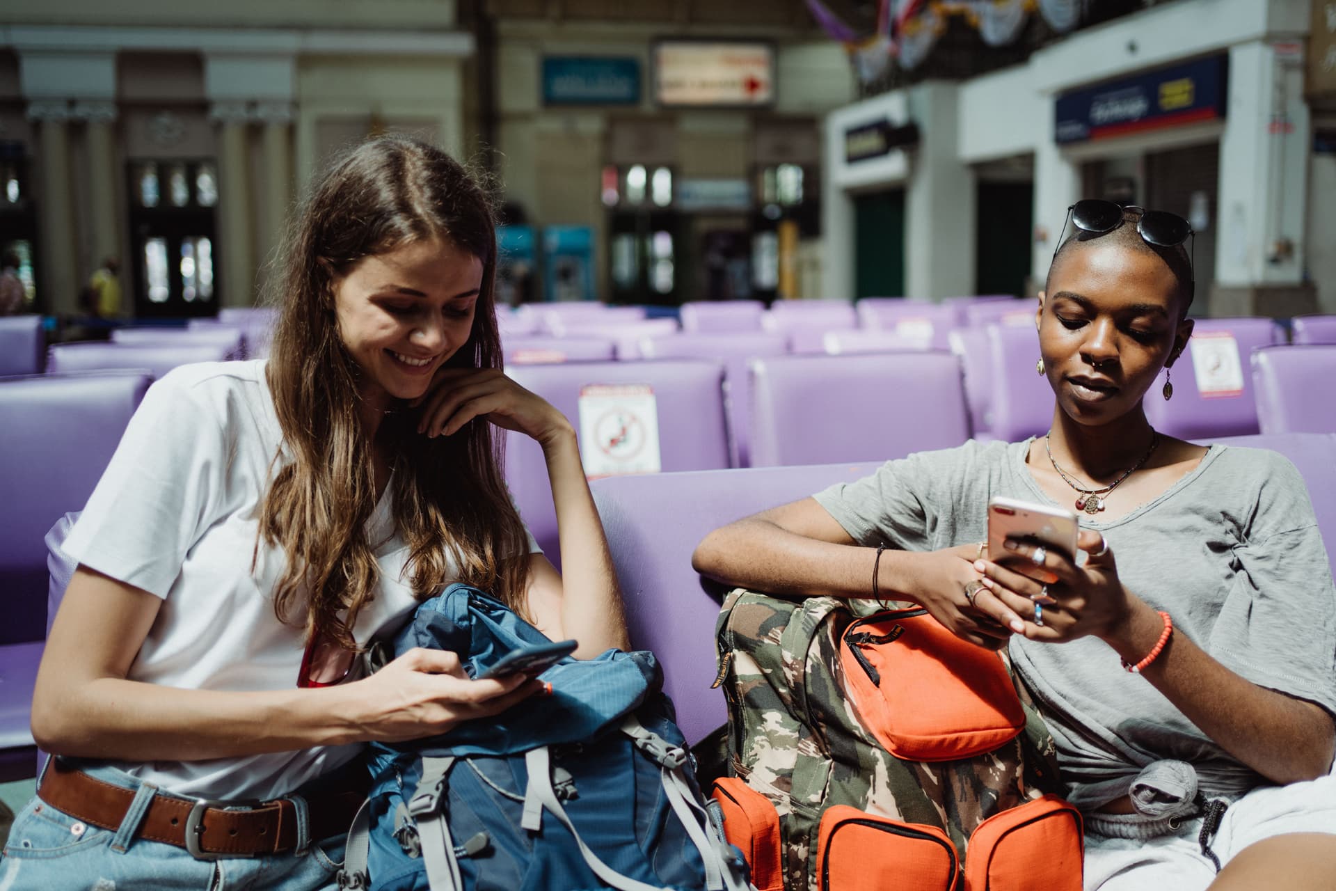 Deux jeunes voyageuses assises dans une salle d'attente de gare, chacune regarde son smartphone et consulte son mobile wallet ou une application de fidélité.