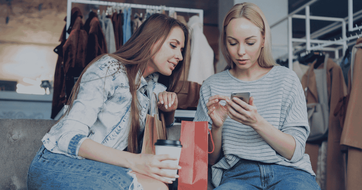 Deux clientes regardent un smartphone dans un magasin de vêtements pour consulter les avantages de leur logiciel de carte de fidélité digitale.